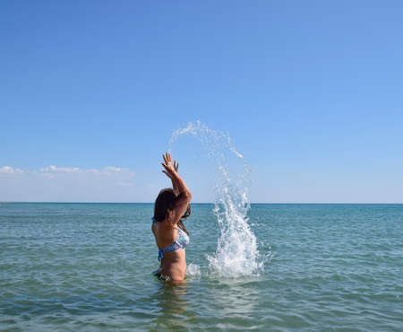 She sprinkles in seawater. Flying up spray from the water throws. The girl with dark hair and a bathing enjoys sea.の写真素材
