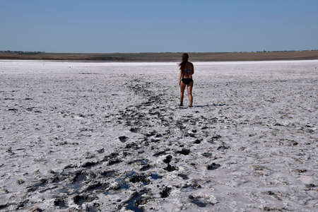 Woman walking along the dry bottom of a salt lake, rear view. Walk the dark-haired woman in a swimsuit on the bottom of a dry lake with salt and mud. The ancient dried-up lake.の写真素材
