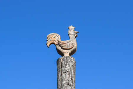 Figurine of a rooster on a tree stump on a background of blue sky. Figures of animals made of wood. Woodcarving.の写真素材