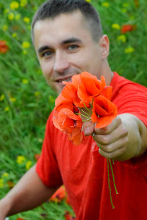 A young man in a red T-shirt is holding a bouquet of red poppies. The man is giving flowers. Poppy field.の写真素材