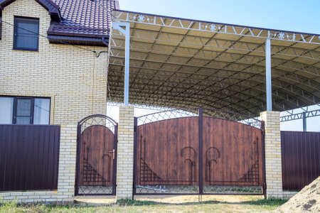 Brick house with a fence and gates. A large awning with a steel frame. View of a new built-up fence and a house made of bricks and corrugated metalの写真素材