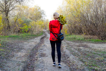 Girl in a red jacket and with a backpack walks along the road to the forest. The beginning of the wayの写真素材