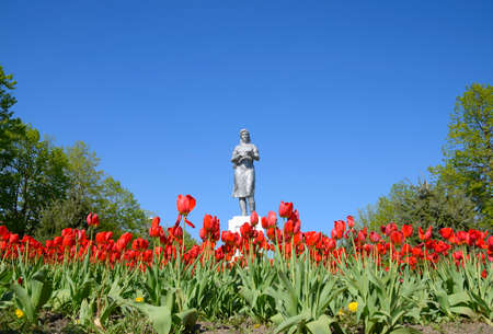 Statue of a collective farmer on a pedestal. The legacy of the Soviet era. A flower bed with tulips and young trees in the village of Oktyabrsky. Krasnodar Krai, Krasnoarmeisky Districtの写真素材