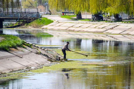 Poltavskaya, Russia - April 07, 2016: An elderly man removes garbage in a dirty city river.のeditorial素材