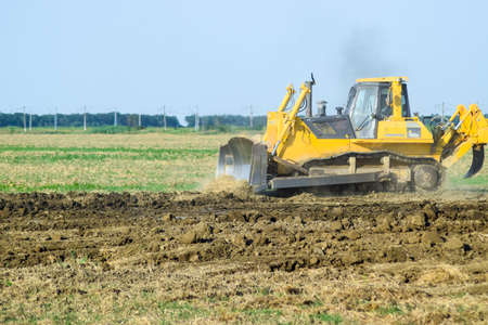 Russia, Temryuk - 15 July 2015: The yellow tractor with attached grederom makes ground leveling. Work on the drainage system in the field.のeditorial素材