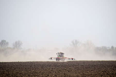 The tractor harrows the soil on the field and creates a cloud of dust behind it.