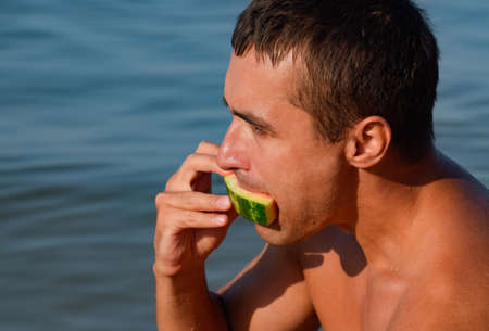 A man is eating a watermelon sitting on the beach. A ripe watermelon in the hands of a man.