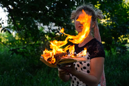 The girl holds a burning book in her hands. A young woman in a forest burns a book.