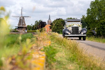 Old bridal car driving through a Dutch landscape with windmill and  church towersの写真素材