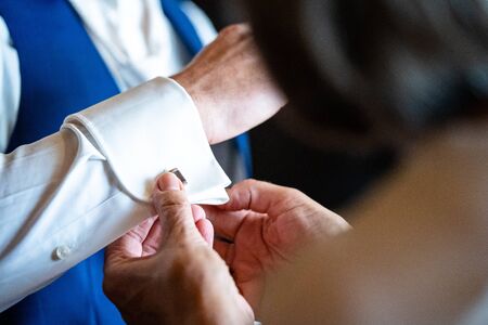 Man in a shirt. Woman helps husband wears a metallic silver cufflinks stud on his arm. Hands groom and bride. Close-up. Wedding day. Morning of the groom.の写真素材