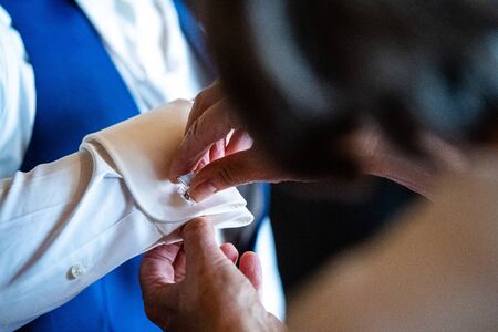 Man in a shirt. Woman helps husband wears a metallic silver cufflinks stud on his arm. Hands groom and bride. Close-up. Wedding day. Morning of the groom.の写真素材