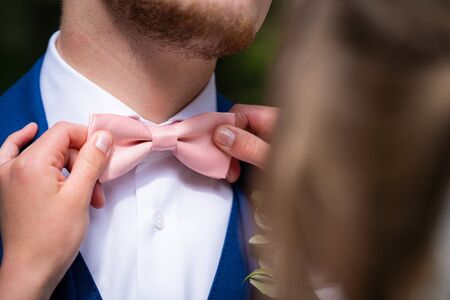 Bow tie in pink color of groom with a blue suit and a white bluze, bride does loving gesture, detail weddingの写真素材