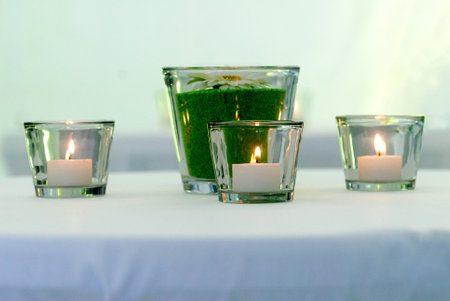 An aged glass candle sits on a table on a white napkin white glass with green and flower in the backgroundの写真素材