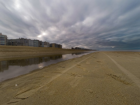 A scenic beach landscape featuring a reflective water body, adjacent modern buildings, and dramatic overcast clouds. Captures the tranquility and serenity of coastal living with a touch of dramatic weather.の写真素材