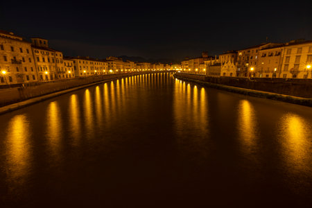 Nighttime view of the Arno River in Pisa, Italy. The scene captures reflections of warm streetlights shimmering along the water.の写真素材