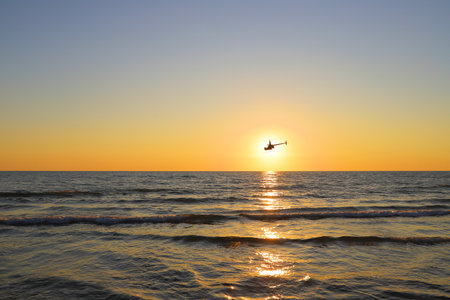 A helicopter's silhouette is visible against a golden-orange sunset, flying over a calm sea. The sun is low on the horizon, creating a reflective path of light on the water.の写真素材