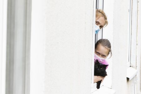 Mom and daughter in medical masks, together with their pet, look out of the window during a pandemic.の写真素材
