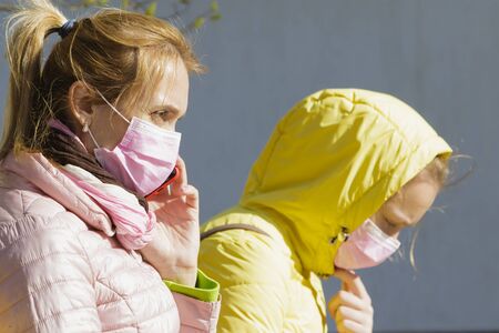 Girl with mom in medical masks sit near the house.