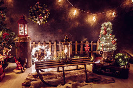 Christmas theme arrangement in dark evening home room. Lot of party string lights illuminated, lanterns, old retro sleight, tiny Christmas tree with dry orange slices, old suitcase, goatskin rug.の写真素材