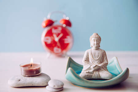 Finding time for self care and balance between body soul and mind. Balance between work and home concept. Lotus pose white color sitting meditation Buddha figurine, red alarm clock on background.の写真素材