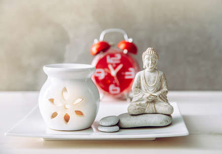 How to find balance between busy life and healthy mental health. Taking a break to sit and meditate concept. Buddha figure with aroma lamp and clock on the background.の写真素材