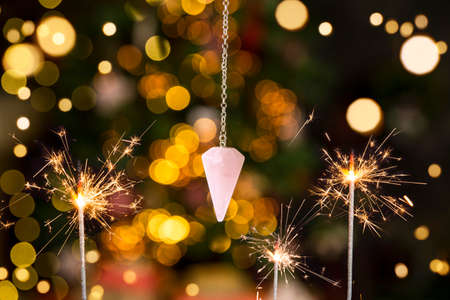 Pink color love rose quartz natural crystal pendulum hanging on silver chain indoors with round yellow bokeh lights on background. New years predictions concept. Room for text.の写真素材