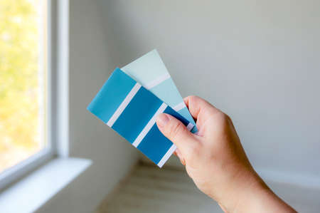 Close up view of person hand holding different blue color scheme and choosing a home wall paint, blurred white empty room background. Home improvements concept.の写真素材