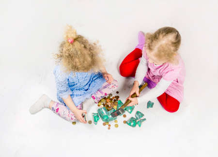 Top view of two young blond girls sisters sit on white floor braking a ceramic piggy bank. Broken piggy bank on floor, euro coins scattered around. Children collecting money and using savings concept.の写真素材