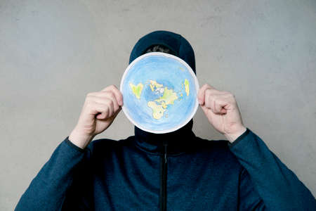 Flat Earther concept. Person who believes that Earth is flat disc. Anonymous hooded Man holding flat Earth model in front of face, copy space. Isolated on gray background, studio shot.の写真素材