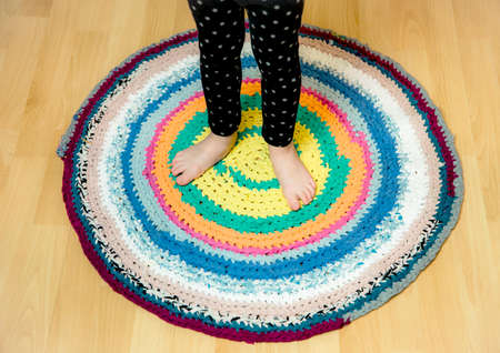 3 year old young child standing on round colorful crochet rug from repurposed T-shirts on brown wooden home floor. Top above view.の写真素材