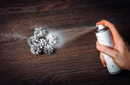 Close up view of person hand using metal canned spray snow to spray decorate Christmas decoration pine cones in winter, art. Studio shot with studio lights indoors dark brown wooden background.の写真素材