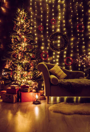 Festive decorated home interior living room in with lot of string LED lights, led curtains and pine cone and berry wreaths on wall, red Christmas gifts under the tree, comfortable canape sofa waiting.の写真素材