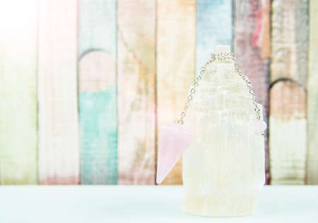 Using crystal Selenite tower for loading rose quartz pendulum, light blue table and pastel color wooden board background. Divination and fortune telling concept.の写真素材