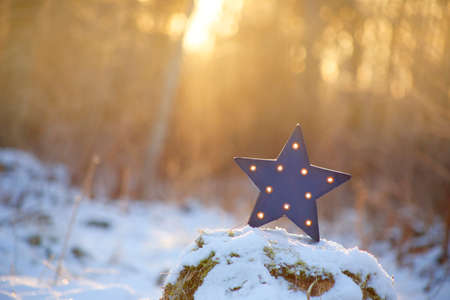 Focus on decorative modern blue star shape lamp with lots of burning small light bulbs on, outdoors on snowy ground, with nice back light from sun setting.の写真素材