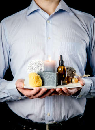 Man holding white ceramic tray with group of different spa essentials, beard oil, gray soap, natural sea sponge, candle, aroma oil black background. Father or husband gift idea concept.の写真素材
