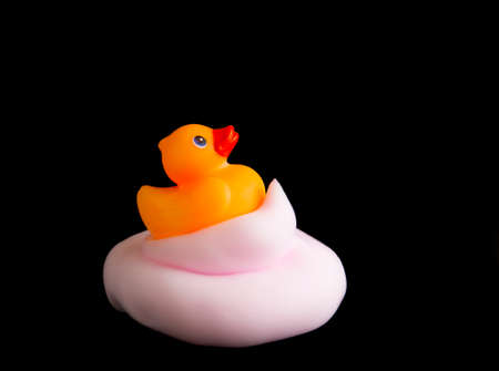Orange rubber bath duck on pile of pink foam isolated on black background.の写真素材