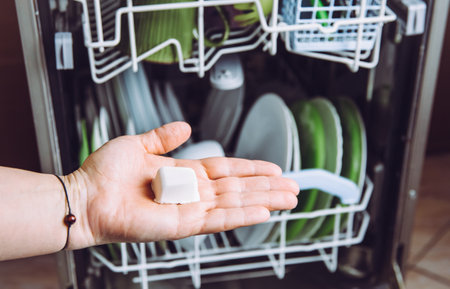 Selective focus on woman hand, holding homemade natural dishwasher pod defocus dishes in dishwasher on background. Green sustainable lifestyle concept.の写真素材