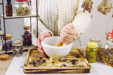 Close up view of woman herbalist mixing various dried herbs for traditional medicinal tea with mortar and pestle. Dried herbs in glass jars on background.の写真素材