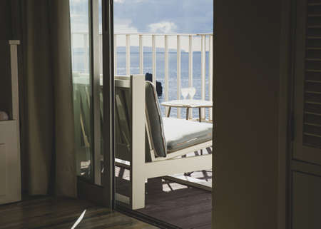 View out of the glass balcony sliding door. White garden chair with blue pillow and table with two glasses with cooling beverage drinks.の写真素材