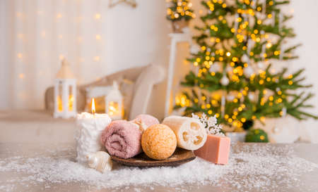 Winter spa pampering concept. Various self care products bath bomb, soap bar, luffa sponge on wood tray with candle burning and Christmas tree with lanterns on background.の写真素材