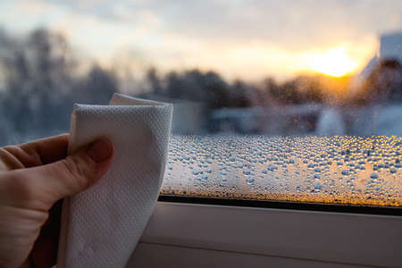 Close up view of person hand using paper cloth, drying wet condensation drops from glass window in...