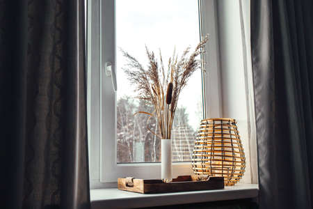 Using dried natural various grass plants inside vase indoors for home decoration. Vase with various plants and wood black metal wire lantern on window sill in daylight.の写真素材
