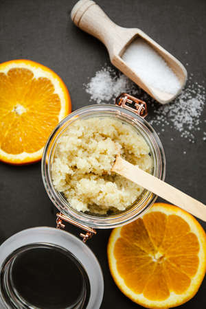 Homemade sugar body scrub in glass jar, with fresh orange. slices and wooden spoon with sugar powder on black stone cutting board. Body skin care concept.の写真素材