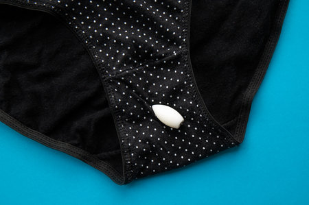 Medicinal bullet shape melting capsule for vaginal use on black dotted underpants, blue indoors background. Moisturizing treatment or infection concept.の写真素材