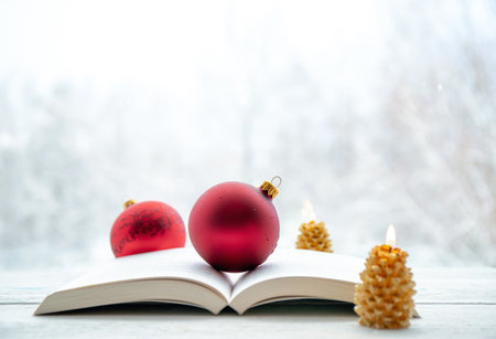 Open paper book with red traditional Christmas baubles and candles burning. Blurred winter landscape on background. Lot of copy space.の写真素材