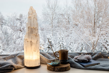 Cozy winter home set with natural selenite crystal electric tower lamp illuminated on window sill, gray color scarf and gloves, paper note book,  tea cup and incense stick. Idyllicの写真素材