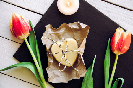 Homemade solid moisturizing hand cream. Heart shape cream bar made of all natural ingredients. Black stone tray background with fresh tulips for decoration spa candle burning.の写真素材