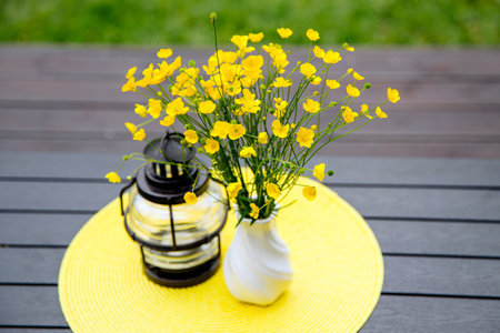 Bouquet of picked yellow Meadow buttercup or Ranunculus acris in a vase on home table, lantern with...
