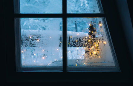 Looking out the frosty winter window. Christmas lights and snow on background. Cozy holiday concept.の写真素材