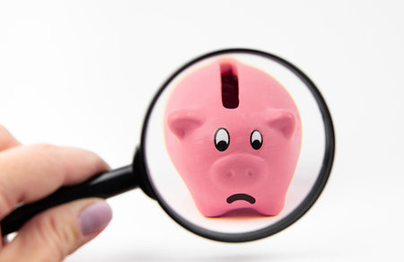 Person hand holding magnifying glass and looking at sad  face piggy bank. Bad finances concept, white studio background.の写真素材